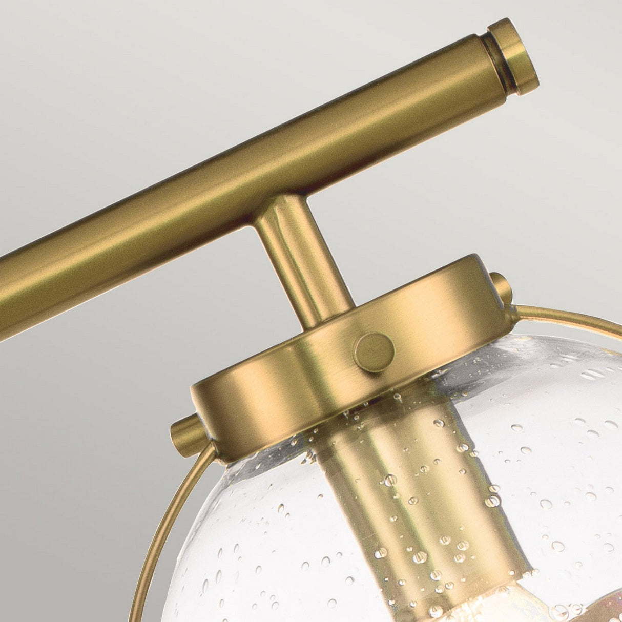Close-up of the Hollis 3 Light Wall Light in Heritage Brass, showcasing its Art Deco-inspired design with a clear glass globe adorned with small bubble-like patterns. The fixture features a sleek, cylindrical holder connected by a horizontal bar, embodying elegance and sophistication.