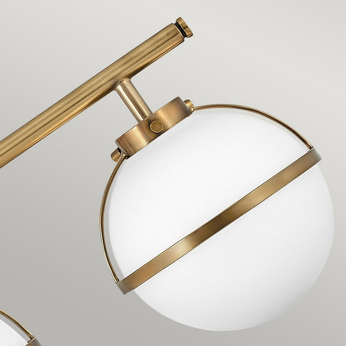 The Hollis 3 Light Wall Light in a heritage brass finish showcases an opal glass shade. Its minimalistic design includes a cylindrical brass support connecting to the spherical element, all set against a neutral gray background.