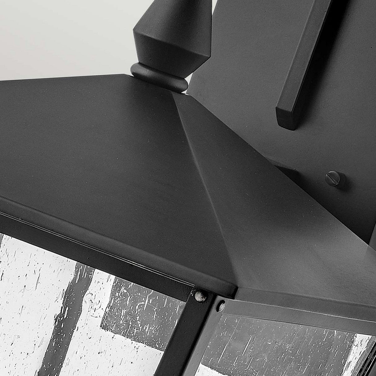 Close-up of the Huntersfield Outdoor 3 Light Large Wall Lantern - Black, featuring a pointed top and clear glass panels. The matte black metal casts intricate reflections of light and shadows, embodying traditional aesthetics. This lantern is perfect for outdoor decor, elegantly mounted on a wall with a visible bracket.