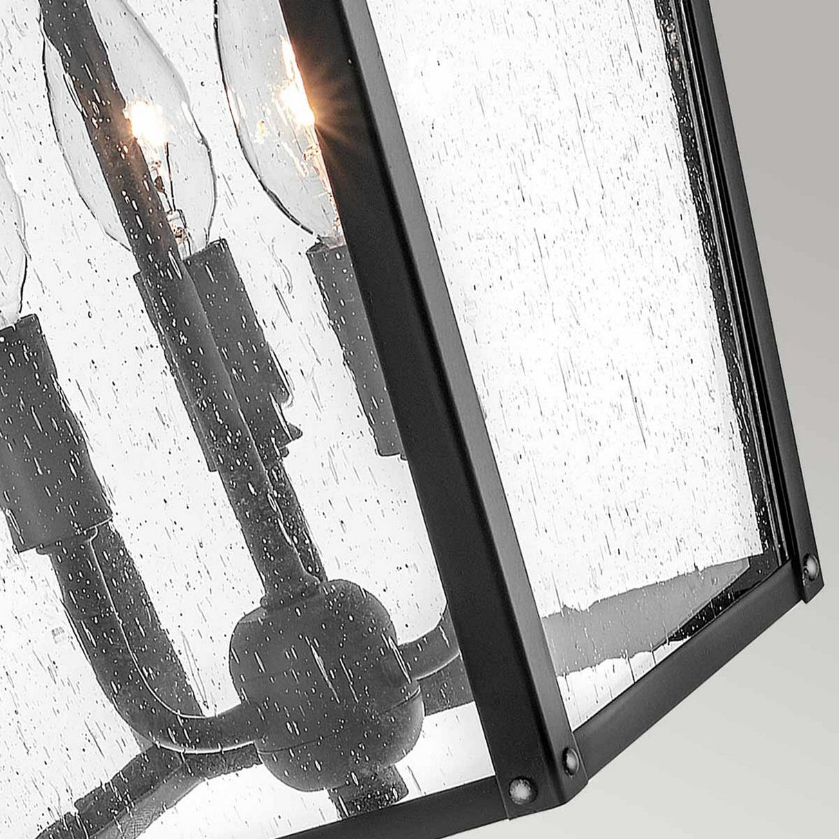 This close-up of the Huntersfield Outdoor 3 Light Large Wall Lantern - Black highlights its rectangular, black-framed design with four candle-like bulbs. The clear, rain-dappled glass adds to its traditional charm, making it a captivating outdoor decor piece.