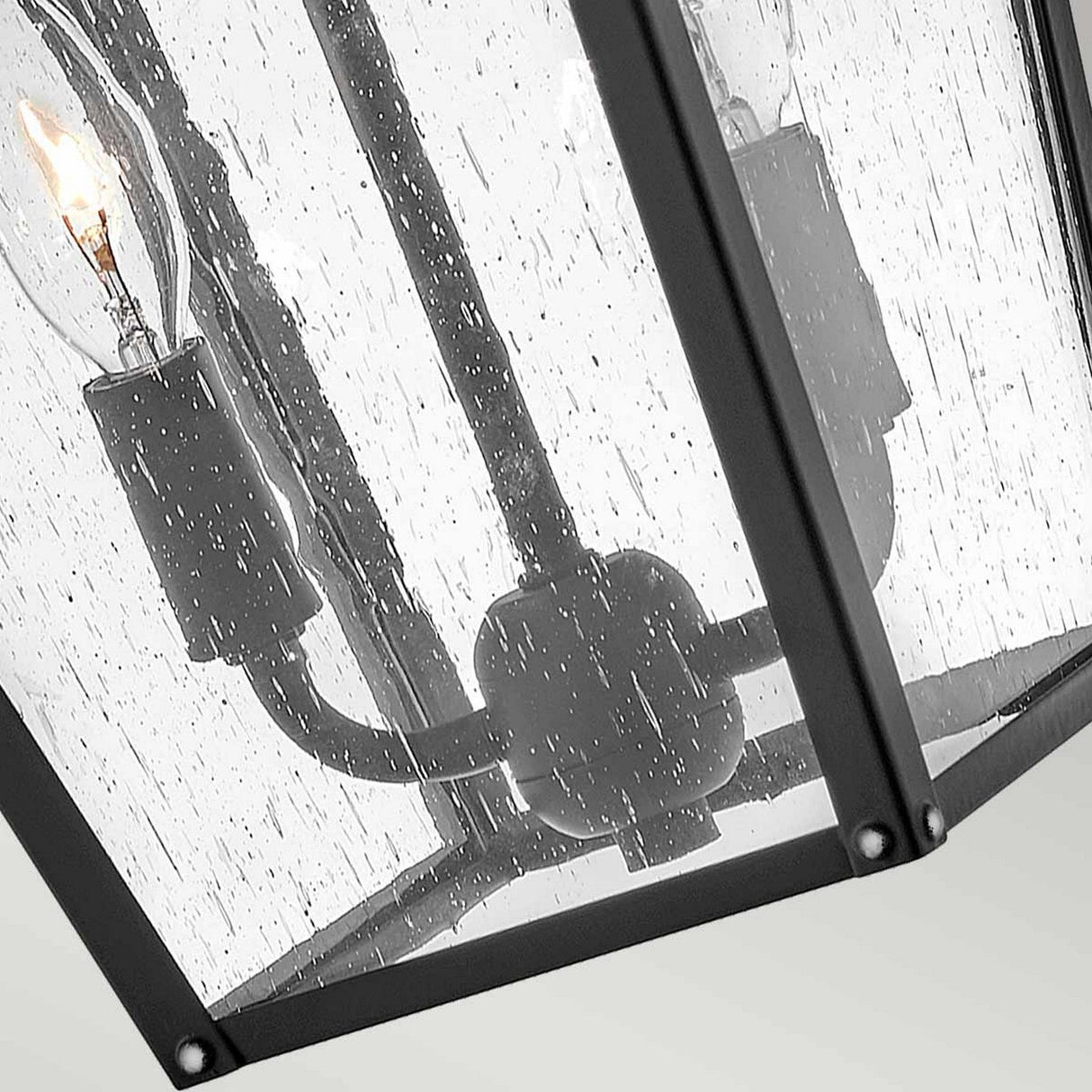 Introducing the Huntersfield Outdoor 2 Light Medium Wall Lantern in sleek black, featuring clear glass panels adorned with a raindrop pattern. This elegant wall lantern houses two candle-style bulbs that glow warmly, supported by matching black metal fixtures, making it ideal for sophisticated outdoor lighting.