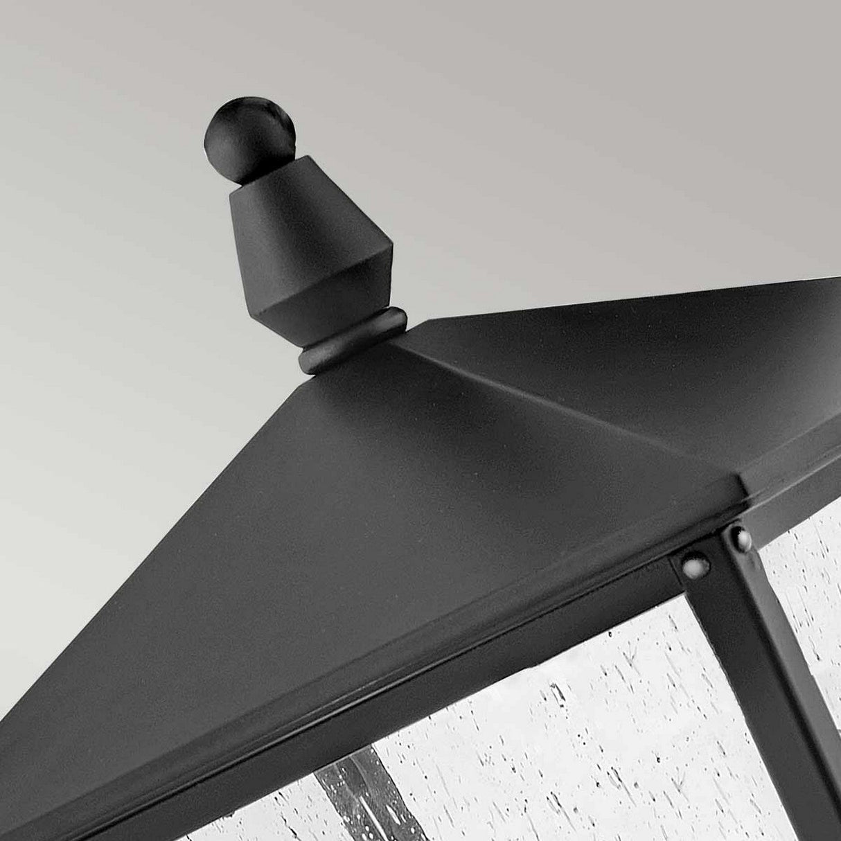 The Huntersfield Outdoor 2 Light Medium Pedestal Lantern in black is featured close up, showcasing its IP44 weather-resistant rating. It has a pointed top finial and clear glass panels with a textured surface, emphasizing its modern design against a light gray background.