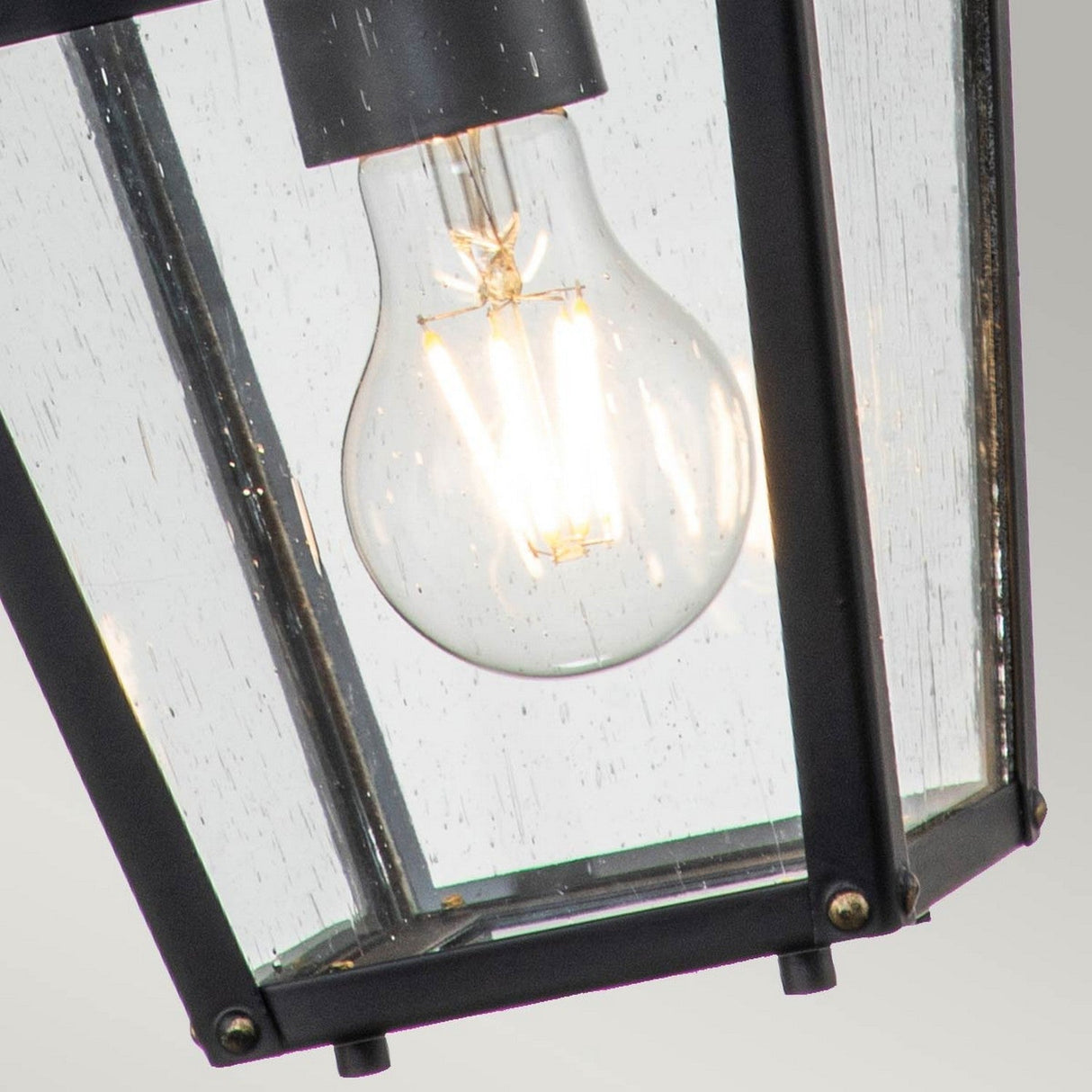 A close-up of the Huntersfield Outdoor 1 Light Small Chain Pendant in black shows a vintage-style bulb inside its metal and glass fixture, featuring clear seeded glass. Raindrops are visible, enhancing the traditional charm as the glowing filament illuminates against the backdrop.