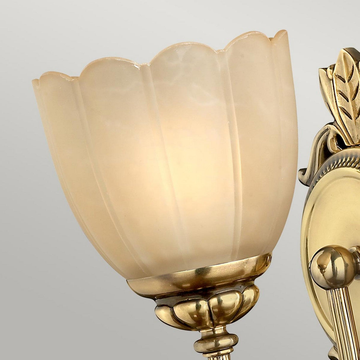 The Isabella 1 Light Wall Light features an etched amber glass shade that casts a warm glow. It is designed with a burnished brass finish, complemented by ornate detailing and a leaf motif on the base, set against a light gray background.
