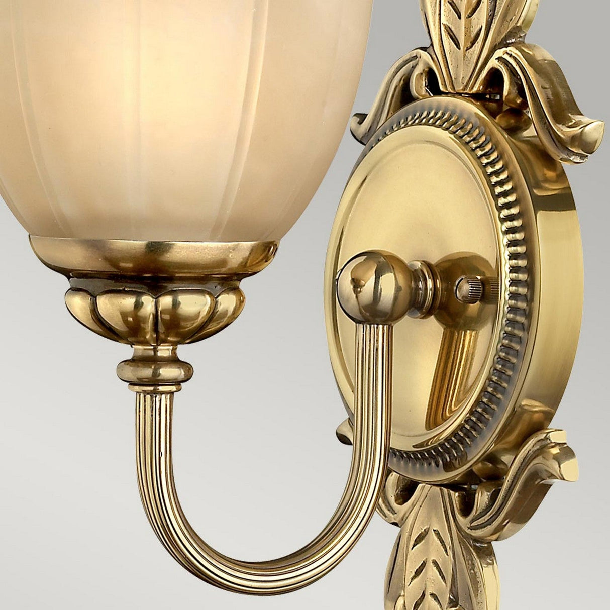 The Isabella 1 Light Wall Light in burnished brass showcases an ornate design with a graceful curved arm, complemented by a marbled etched amber glass shade that emits a warm, soft glow. Set against a simple gray background, the wall light's intricate craftsmanship is beautifully highlighted.
