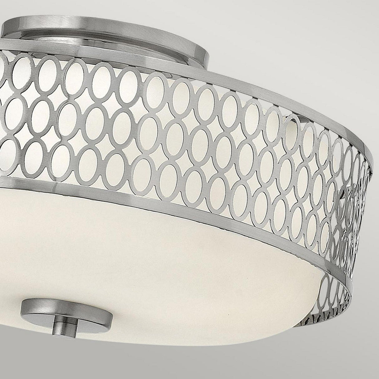 The Jules 3 Light Flush - Brushed Nickel features a circular metal frame with an intricate oval pattern and is complemented by a white glass diffuser, giving it a modern and elegant appearance.