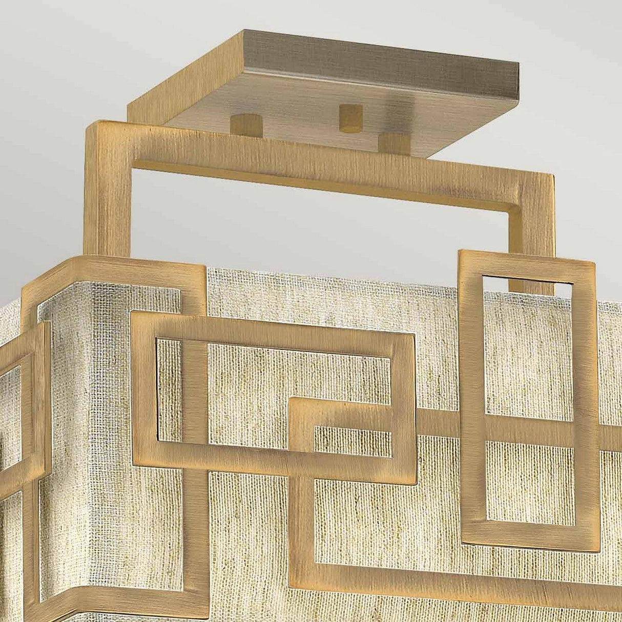 A detailed view of the Lanza 3 Light Semi Flush - Brushed Bronze ceiling fixture highlights its geometric design. It has interlocking rectangular shapes with a brushed bronze finish, surrounding a textured, fabric-like diffuser and is stylishly mounted on a square ceiling plate.