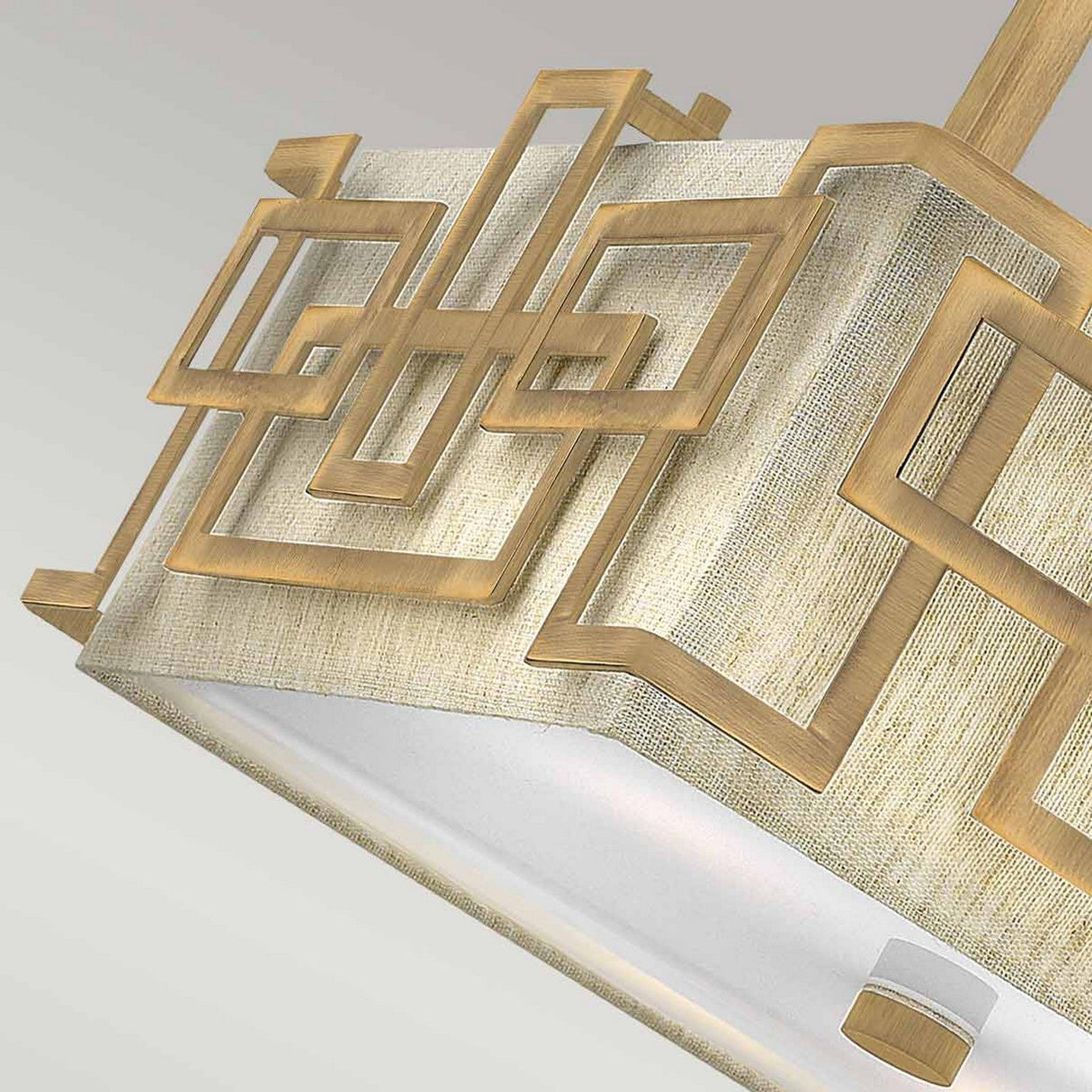 A close-up of the Lanza 3 Light Semi Flush in Brushed Bronze highlights its decorative geometric design with overlapping squares. Its lampshade, made from textured beige fabric, is set against a gradient background transitioning from light gray to white.