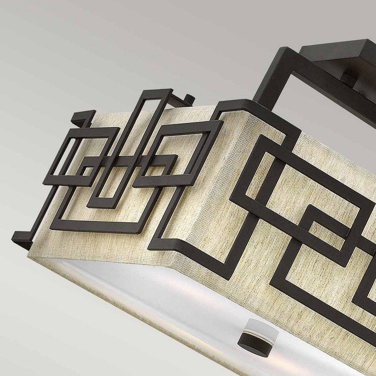 Close-up of the Lanza 3 Light Semi Flush - Oil Rubbed Bronze ceiling fixture, featuring a rectangular beige fabric shade adorned with complex dark geometric metal designs in oil-rubbed bronze for a sleek, modern look. The background showcases a simple gradient.