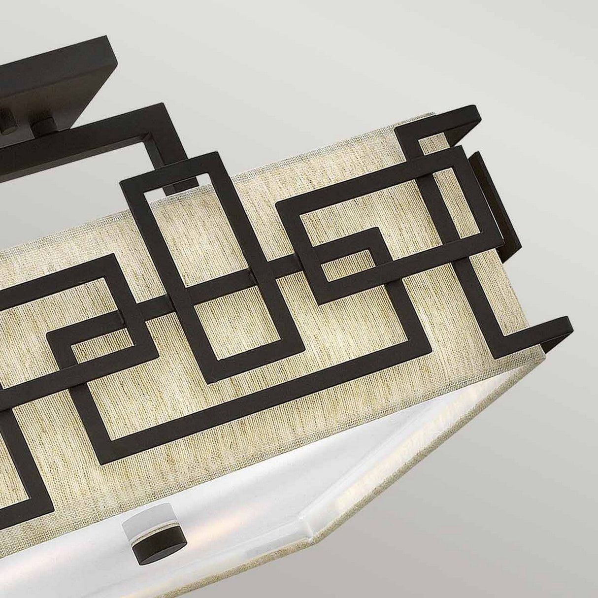 A close-up of the Lanza 3 Light Semi Flush in Oil Rubbed Bronze highlights its contemporary design, featuring a metal frame with geometric patterns overlaying a beige fabric shade against a neutral gray wall.