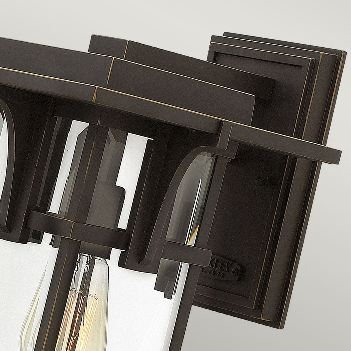 A detailed view of the Manhattan Outdoor 1 Light Medium Wall Lantern showcases its modern design with an Oil Rubbed Bronze finish and clear glass panels. The lantern features clean lines, an exposed bulb, and is mounted on a square backplate against a light gray background. Its IP44 rating makes it ideal for outdoor use.