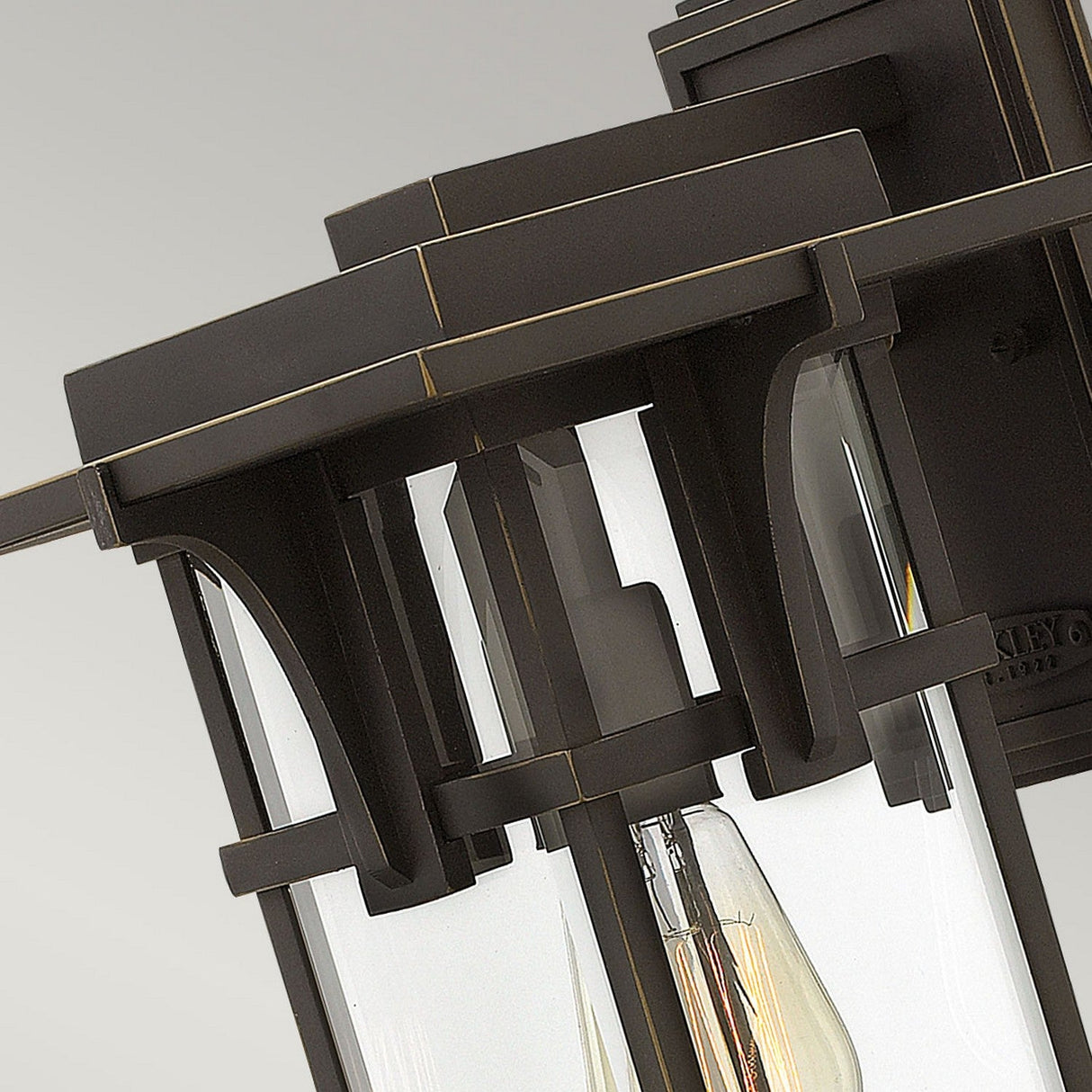 Detailed view of the Manhattan Outdoor 1 Light Medium Wall Lantern in oil rubbed bronze, featuring clear glass panels. This fixture showcases a geometric design with a visible light bulb, offering an industrial aesthetic against a light grey background and boasts an IP44 rating for durability.