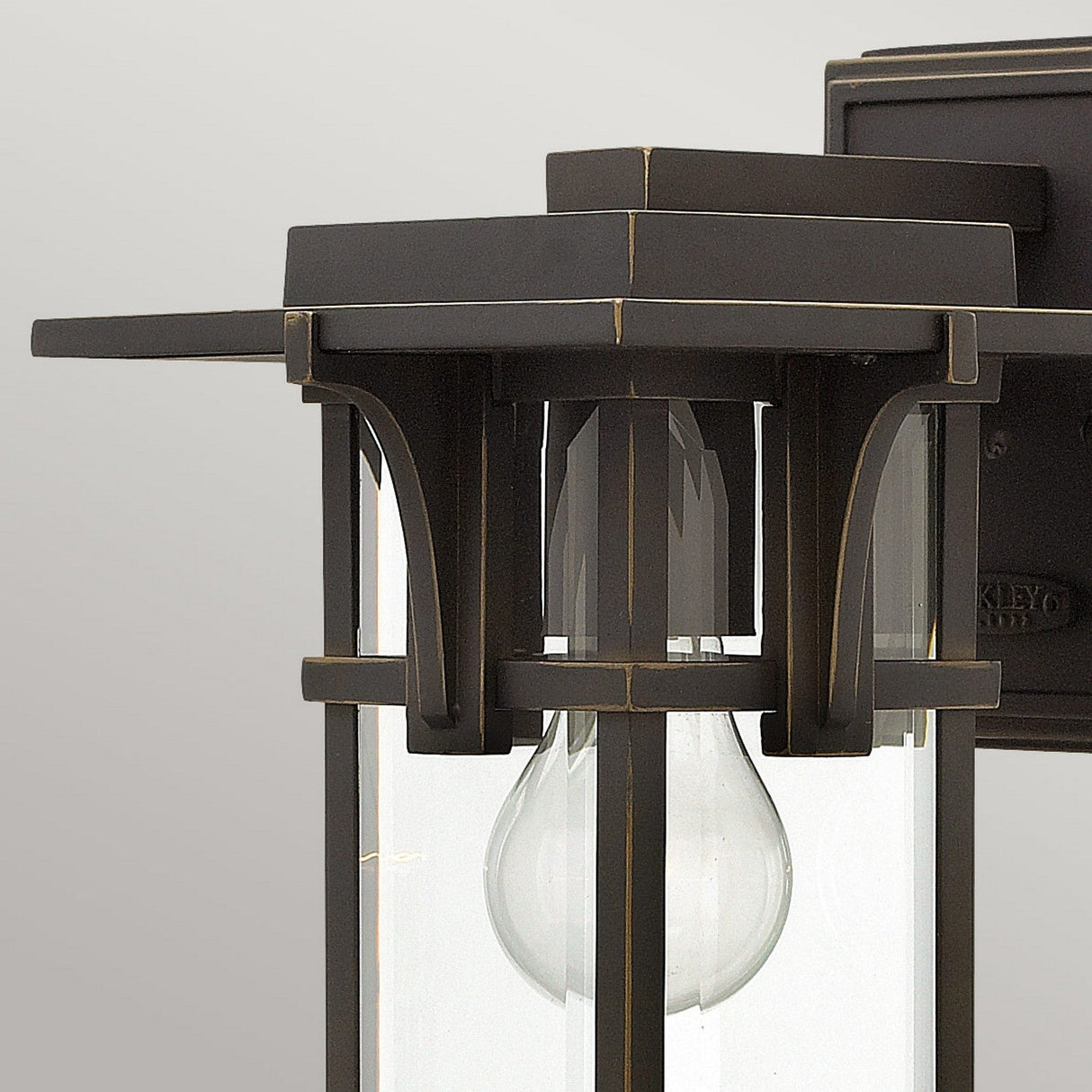 Close-up of the Manhattan Outdoor 1 Light Small Wall Lantern, featuring an oil rubbed bronze finish and clear glass panels. Inside, a bulb is visible. The fixture's clean lines and contemporary design against a plain gray background subtly hint at vintage inspiration.