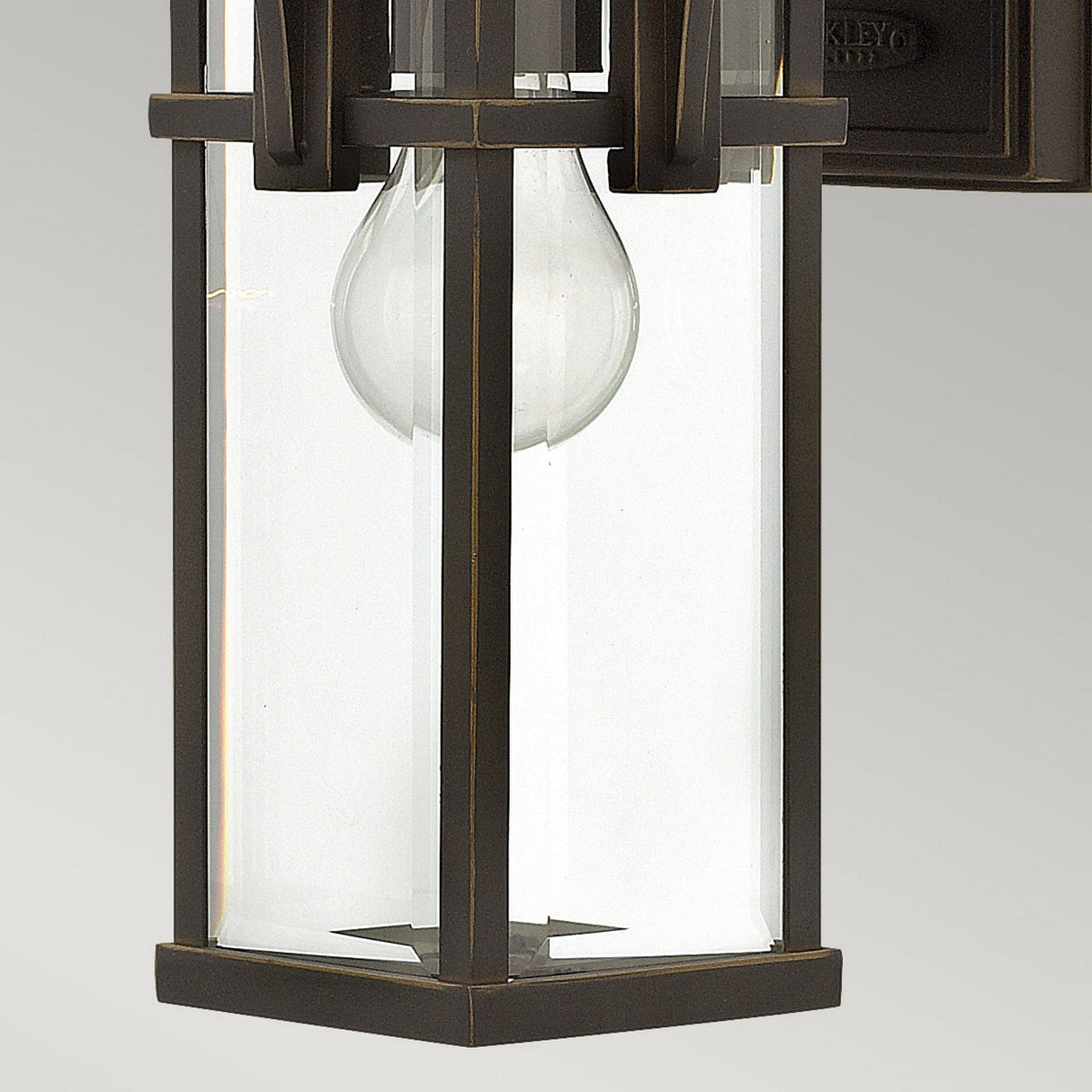 A close-up of the Manhattan Outdoor 1 Light Small Wall Lantern showcases its rectangular shape with a glass casing and visible light bulb. The frame is finished in oil-rubbed bronze and highlights a minimalist design. The plain, light gray background enhances its vintage-inspired look.