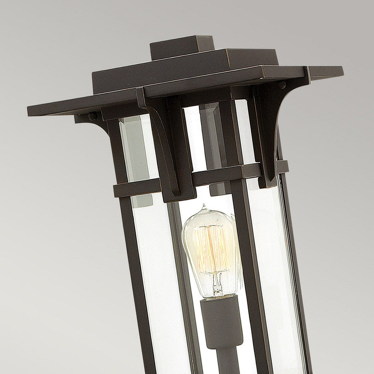 A close-up of the Manhattan Outdoor 1 Light Pedestal in oil rubbed bronze shows its rectangular frame and clear glass panels. Inside, an exposed filament bulb glows warmly against a gray background, combining style with safety thanks to its IP44 rating for all-weather durability.