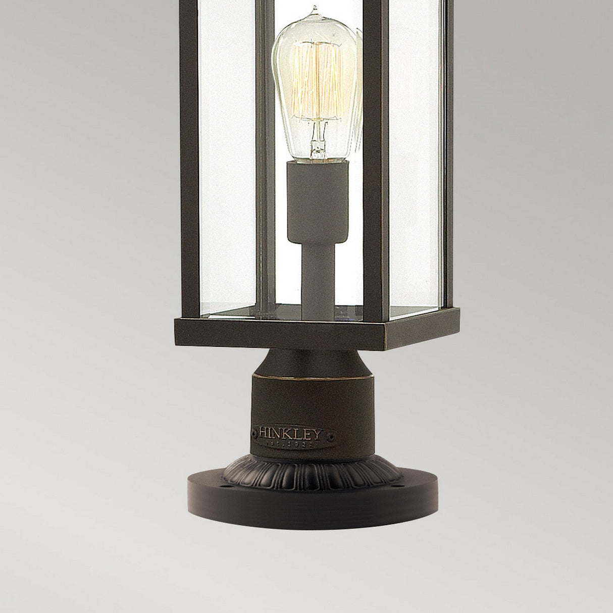 A contemporary outdoor pedestal lantern in oil-rubbed bronze, showcasing a clear glass enclosure with a visible Edison bulb, set against a light gray background. This IP44-rated fixture features intricate detailing on its base and is embossed with the name Hinkley, ensuring both style and durability. It is known as the Manhattan Outdoor 1 Light Pedestal.