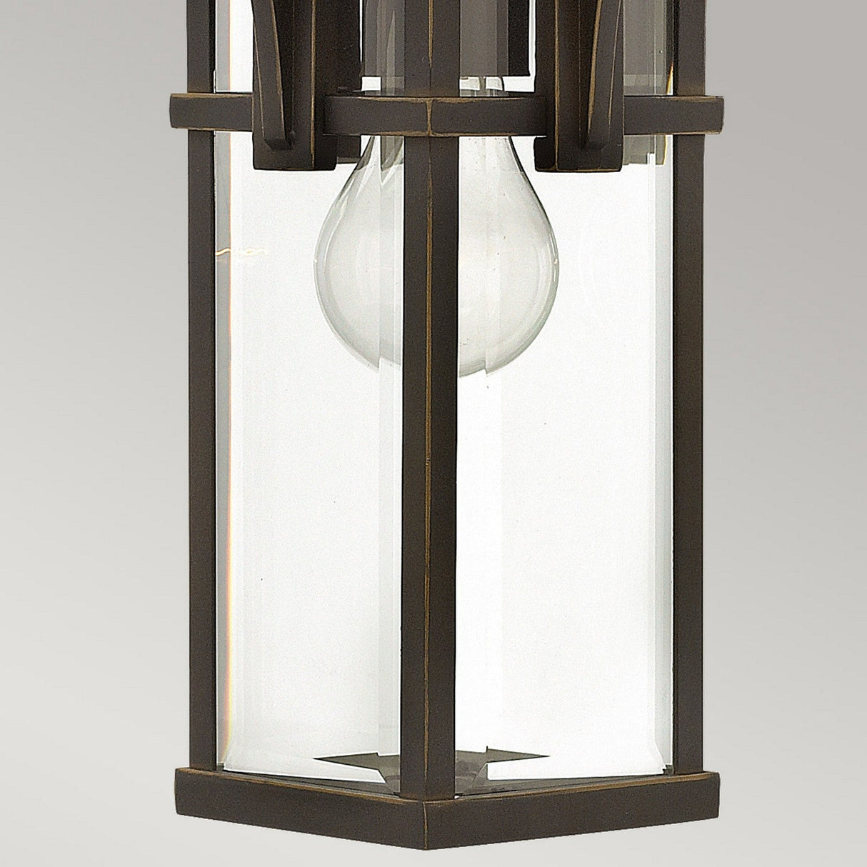 A close-up of the Manhattan Outdoor 1 Light Chain Pendant reveals its hexagonal design with a glass enclosure and visible bulb. This fixture, evoking a vintage train station lantern style, boasts an oil-rubbed bronze finish and is highlighted against a neutral gray backdrop.