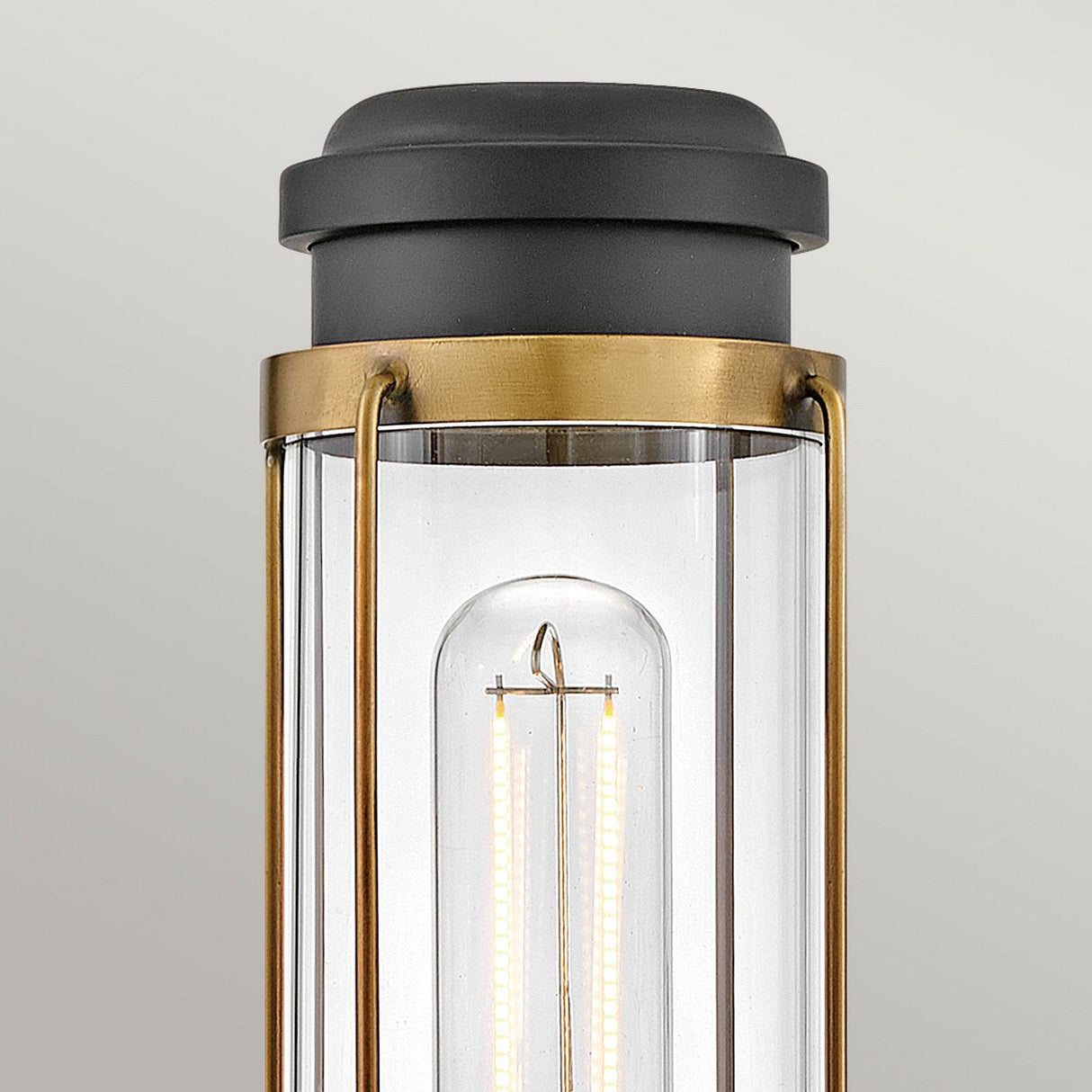 Close-up of the Masthead 1 Light Wall Light - Heritage Brass & Black, featuring a cylindrical glass enclosure that reveals a visible LED filament bulb. The brass finish and black cap enhance its nautical charm against a plain background.