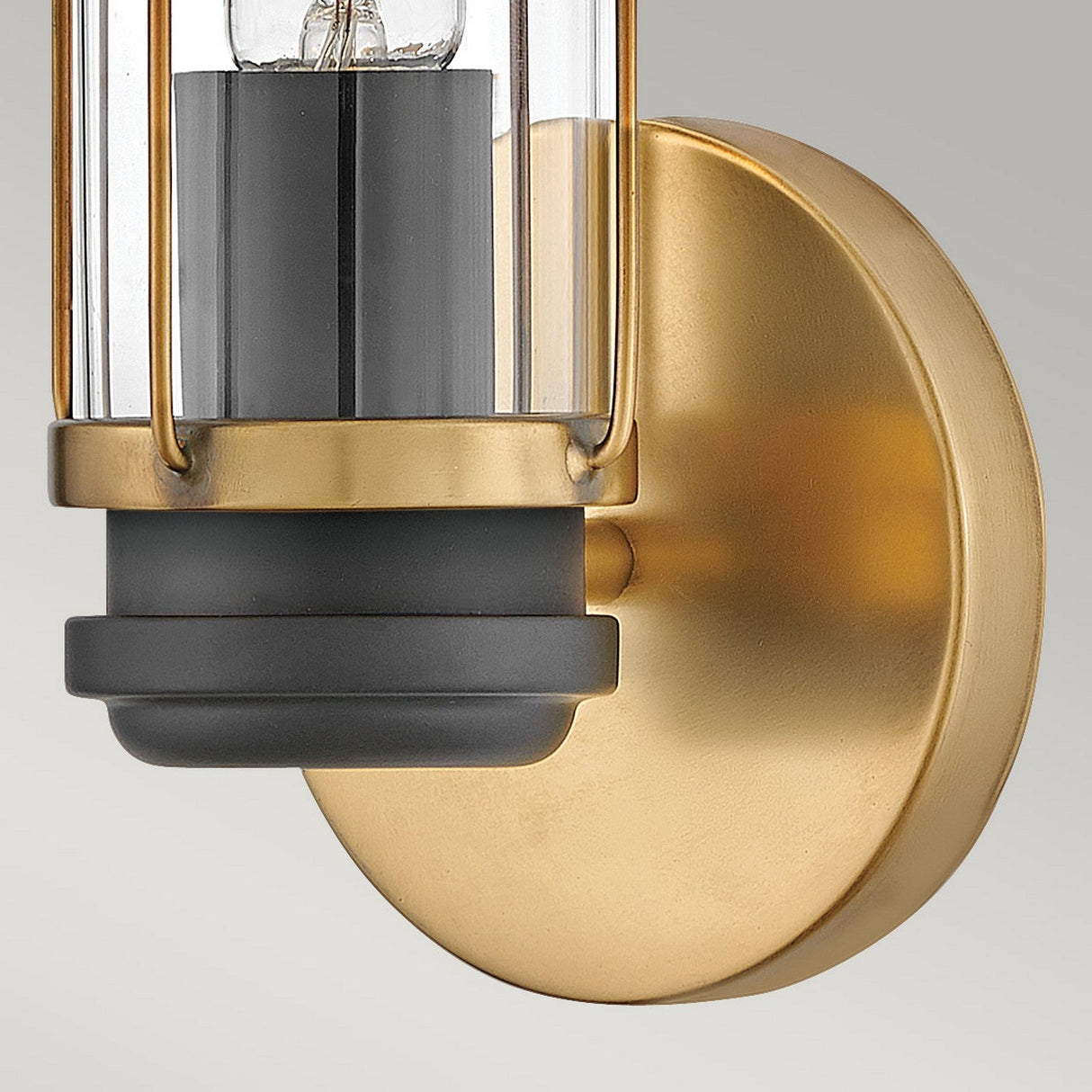 A close-up of the Masthead 1 Light Wall Light in Heritage Brass & Black showcases its nautical allure with a brass circular base and a clear cylindrical glass shade. The visible black socket inside adds an industrial touch, beautifully merging classic and industrial elements to embody a transitional design style.