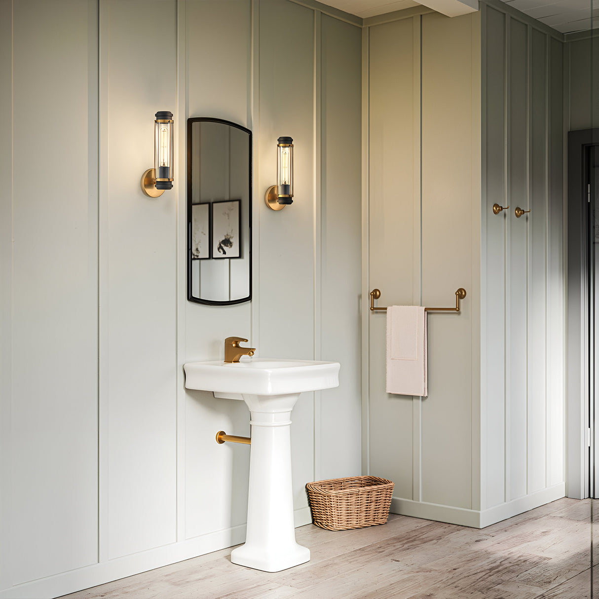 A minimalist bathroom features a white pedestal sink with gold fixtures, topped by a Masthead 1 Light Wall Light in Heritage Brass & Black, which adds nautical charm. A rectangular mirror is mounted on the paneled wall, accompanied by a wicker basket on the floor and a towel elegantly draped over a gleaming gold towel bar.