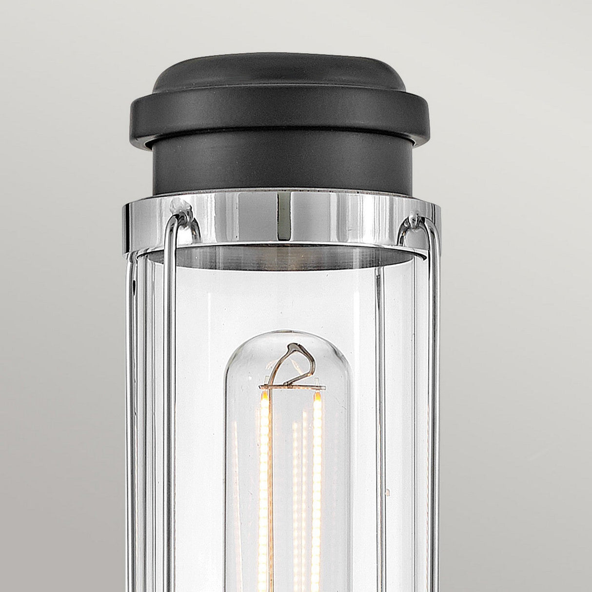 Close-up of the Masthead 1 Light Wall Light - Polished Chrome & Black, showcasing its cylindrical transparent glass casing. The visible LED filament bulb inside shines brightly, while the fixture exudes nautical charm with a sleek black cap and polished chrome accents against a neutral backdrop.