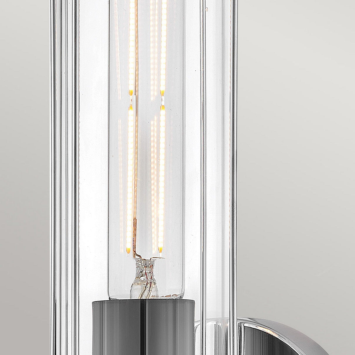 A detailed view of a contemporary light fixture showcases an elongated clear glass bulb with visible filaments, encased in a sleek cylindrical holder crafted from polished chrome. Set against a neutral backdrop, it exudes a subtle nautical allure reminiscent of the Masthead 1 Light Wall Light - Polished Chrome & Black design.
