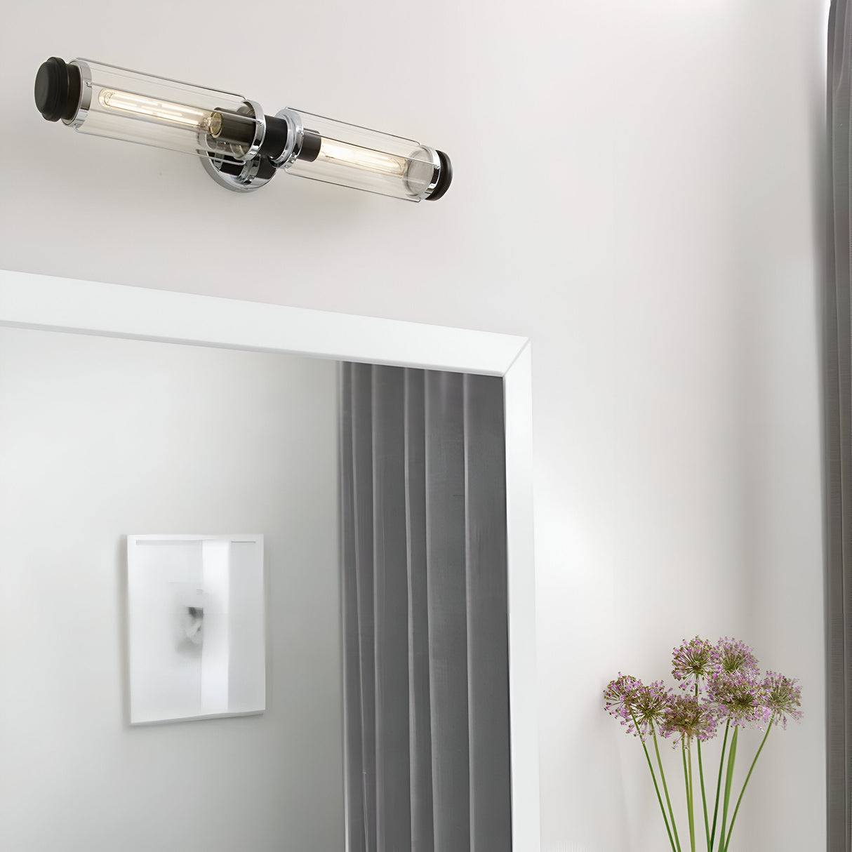 A modern bathroom exudes nautical charm with a rectangular mirror framed in white. Above it, the Masthead 2 Light Wall Light in polished chrome and black showcases exposed bulbs. To the right, purple allium flowers rest in a vase, while soft curtains hang gracefully beside the mirror.