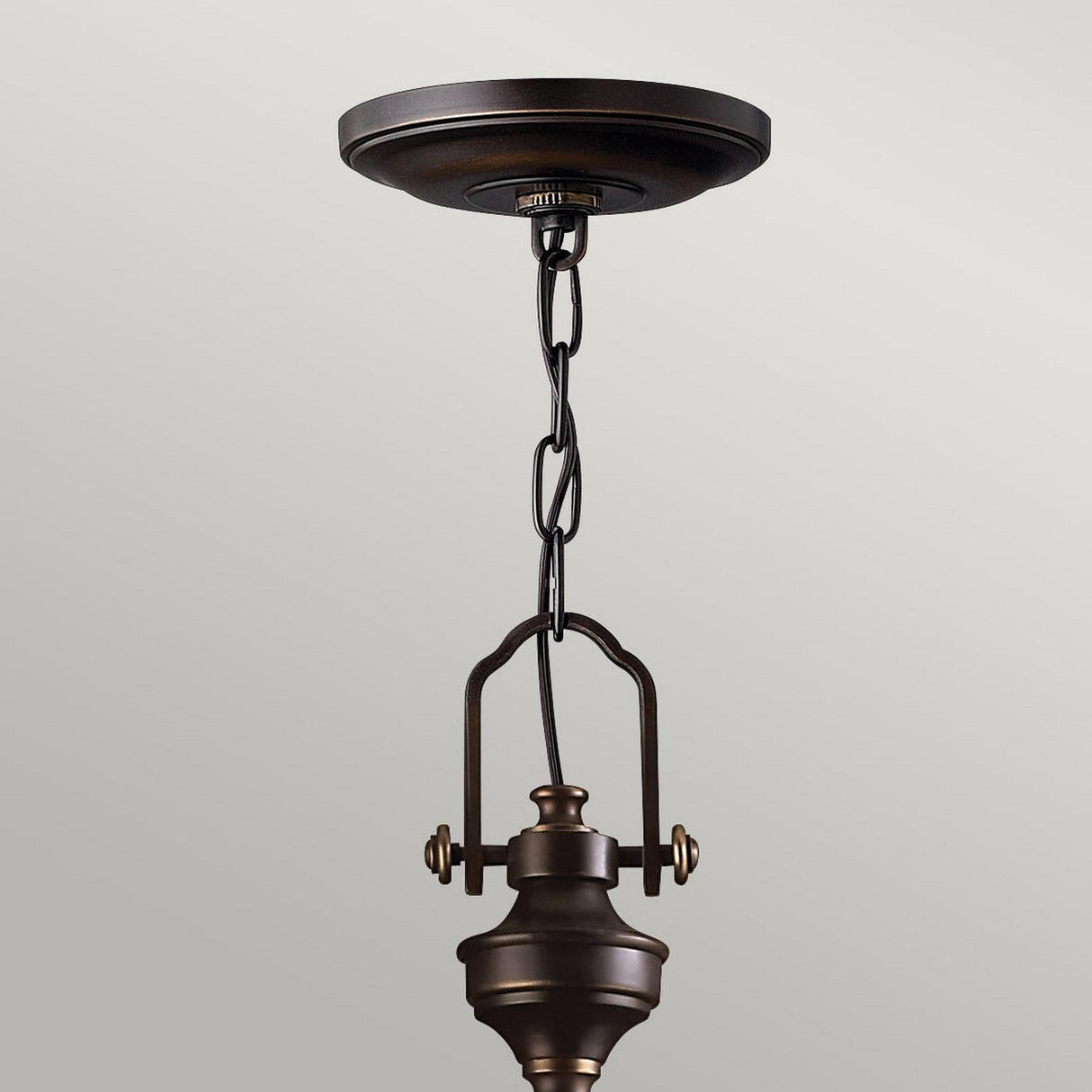 The Mayflower 3 Light Pendant - Bronze gracefully hangs from the ceiling, featuring a decorative chain and minimalist design. Its adjustable height pairs beautifully with the simple, light gray backdrop.