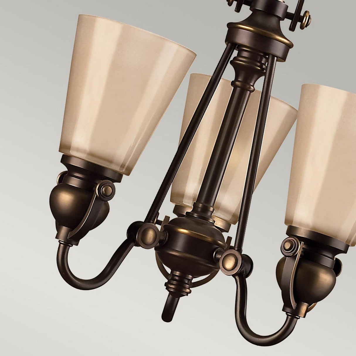 The Mayflower 3 Light Chandelier - Bronze is elegantly showcased in a close-up view, highlighting its bronze finish and three etched amber glass cone-shaped shades. The detailed metalwork of the fixture stands out, emphasizing its traditional elegance against a neutral gray background.