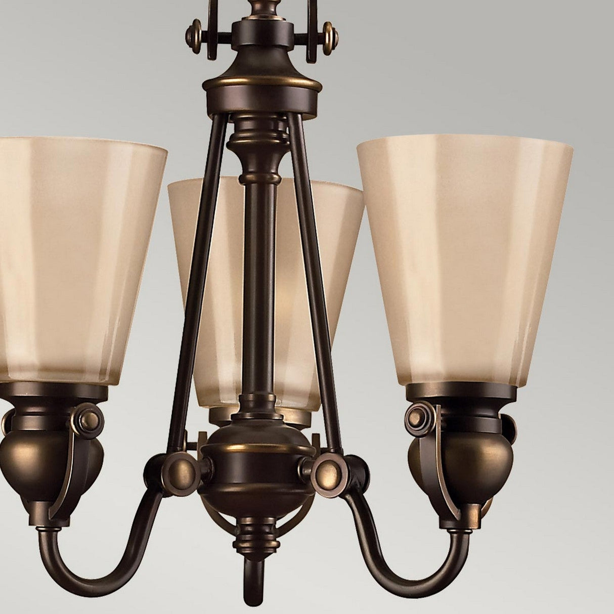 The Mayflower 3 Light Chandelier - Bronze exudes traditional elegance with its sophisticated bronze finish, featuring three upward-facing frosted glass shades. Its exquisite detailed metalwork and central support design are perfectly highlighted against a neutral gray background.