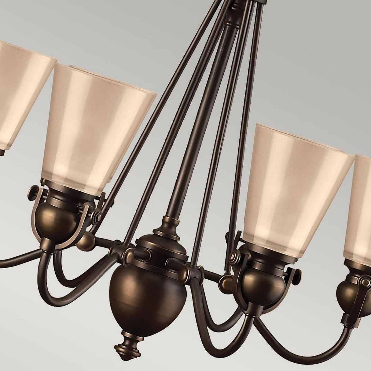 Close-up of the Mayflower 6 Light Chandelier with five elegant, cream-colored, tapered glass shades and an etched amber glass shade. Its bronze finish and gracefully curved arms stand out against a plain gray background.