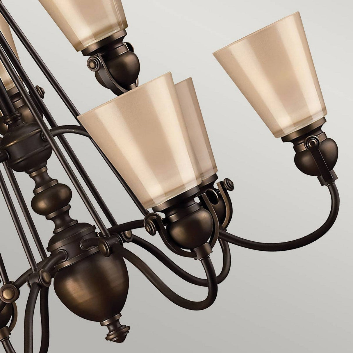 A detailed view of the Mayflower 9 Light Chandelier - Bronze highlights its gracefully curved arms and frosted cone-shaped glass shades, set against a gray background.