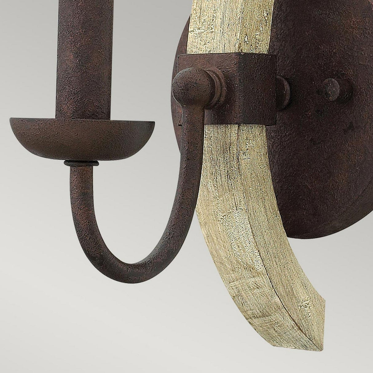 A close-up of the Middlefield 1 Light Wall Light in Iron Rust showcases its rustic elegance with a dark metal frame and gracefully curved beige wooden accent. The minimalist design features a U-shaped arm supporting a small, round platform for the bulb, set against a neutral gray background to enhance its farmhouse lighting charm.