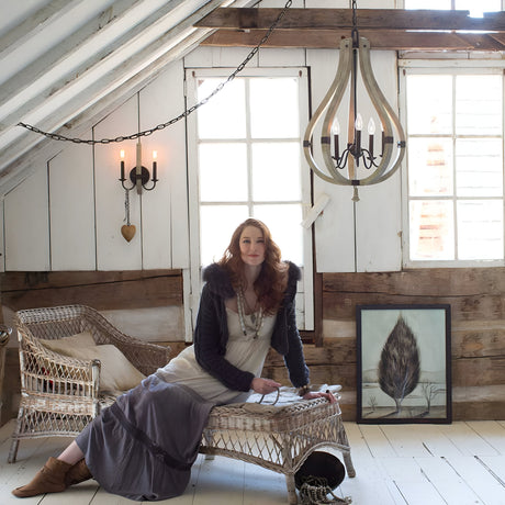 A woman with long red hair relaxes on a wicker chaise in a rustic attic room, exuding charm. She wears a long skirt, blouse, and fur-trimmed cardigan. The room is adorned with wooden beams, large windows, the Middlefield 5 Light Chandelier in Iron Rust hue, and feather artwork on the floor.