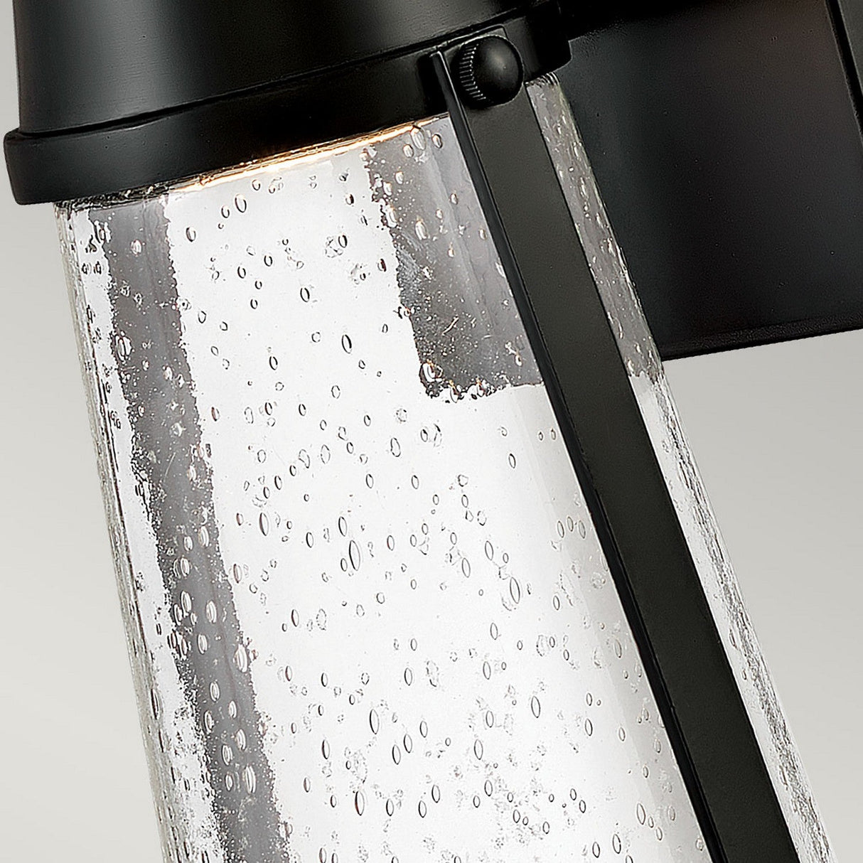 Close-up of the Miles 1 Light Wall Lantern - Black, showcasing its modern design with a textured glass panel filled with numerous small bubbles. The black metal frame contrasts against the clear glass, evoking a subtle nautical theme while a warm, energy-efficient light glows inside the lamp.