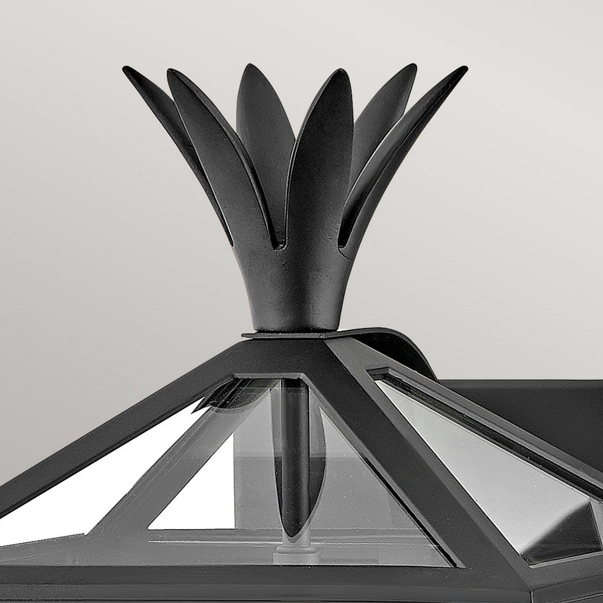 The Palma Outdoor 2 Light Medium Wall Lantern - Black features a decorative black metal ornament with a stylized flower or flame design, crowning a glass and metal structure with triangular glass panels, all set against a plain, light gray background.
