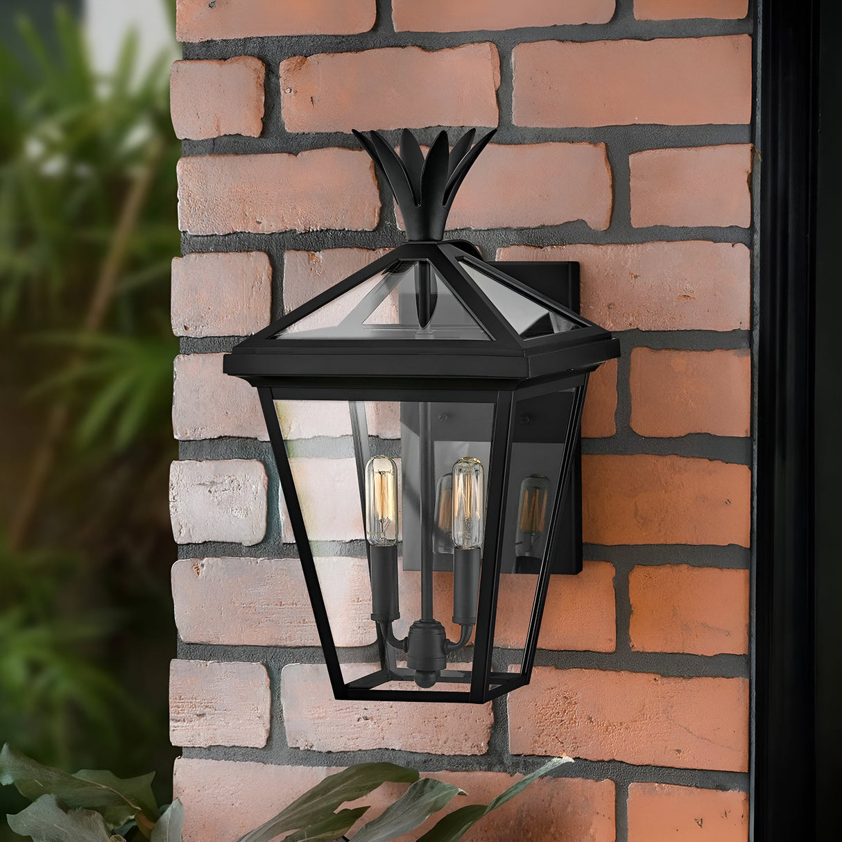 The Palma Outdoor 2 Light Medium Wall Lantern - Black, featuring a decorative pineapple motif, is mounted on a red brick wall. This black metal design includes glass panels that display two visible bulbs, set against a backdrop of lush greenery.