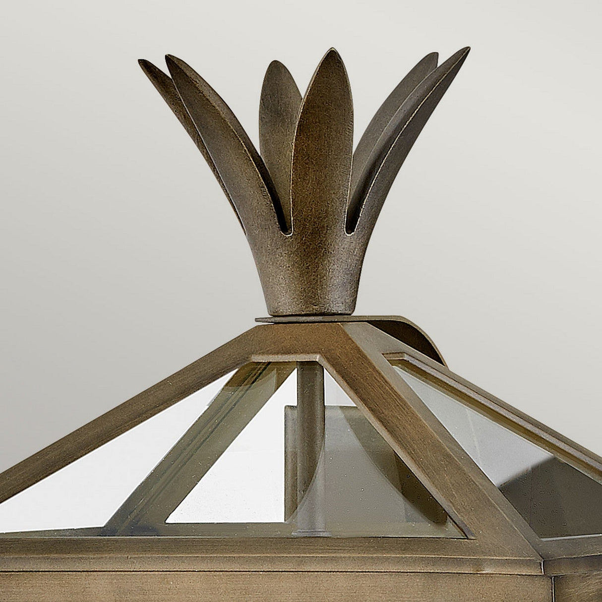 The Palma Outdoor 2 Light Medium Wall Lantern in Burnished Bronze features a metal, petal-like finial atop a geometric structure with glass panels, subtly evoking a pineapple motif. Set against a plain, light background, this lantern is an exquisite piece of outdoor decor.