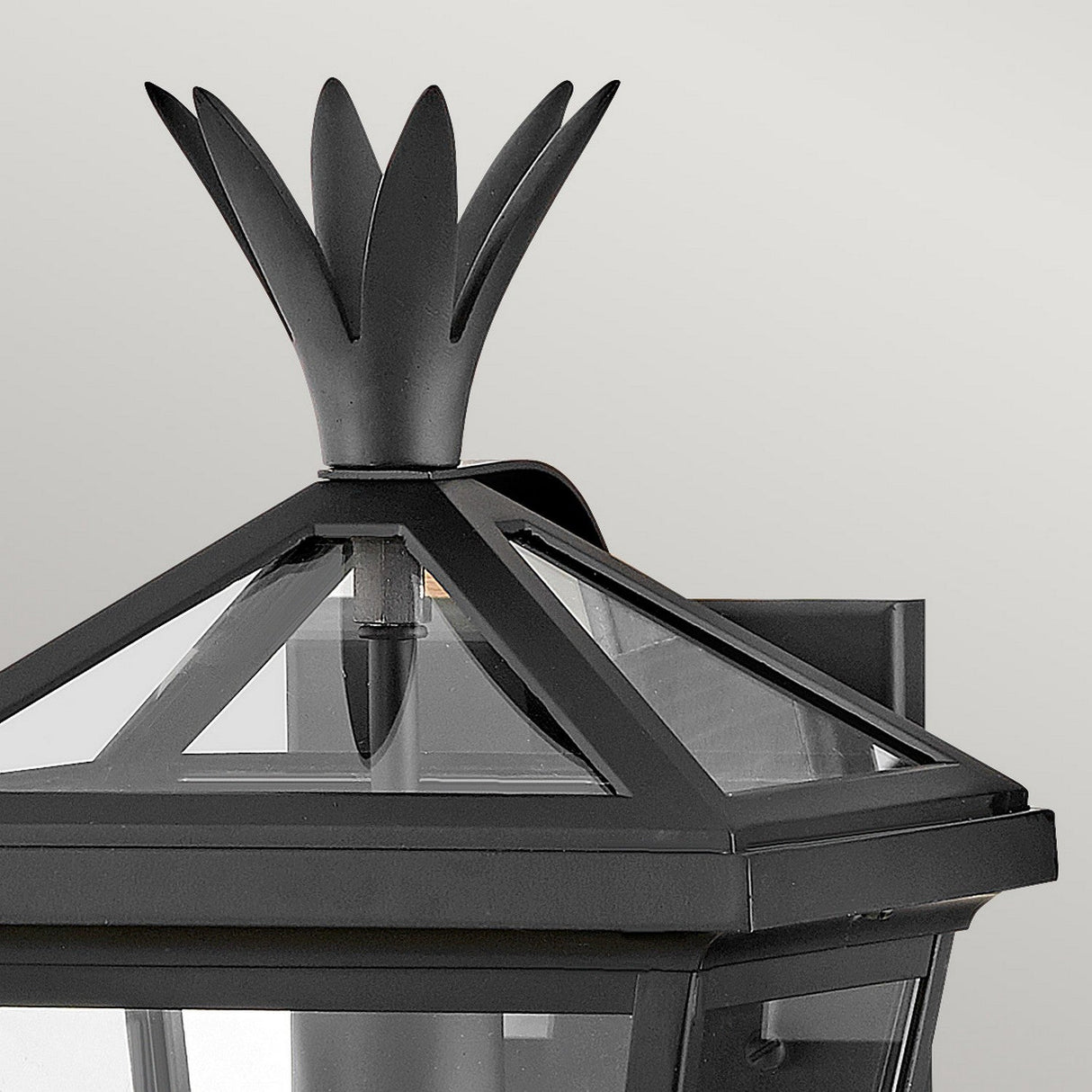 Close-up of the Palma Outdoor 1 Light Small Wall Lantern in black, featuring a geometric design with an ornamental pineapple motif on top and clear glass panels, elegantly contrasting against the neutral light gray background.