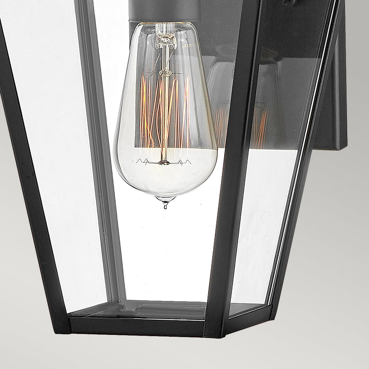 A close-up of the Palma Outdoor 1 Light Small Wall Lantern showcases its modern design, featuring a clear glass enclosure with an exposed filament bulb. The fixture's sleek black finish and clean lines are elegantly mounted against a plain background.