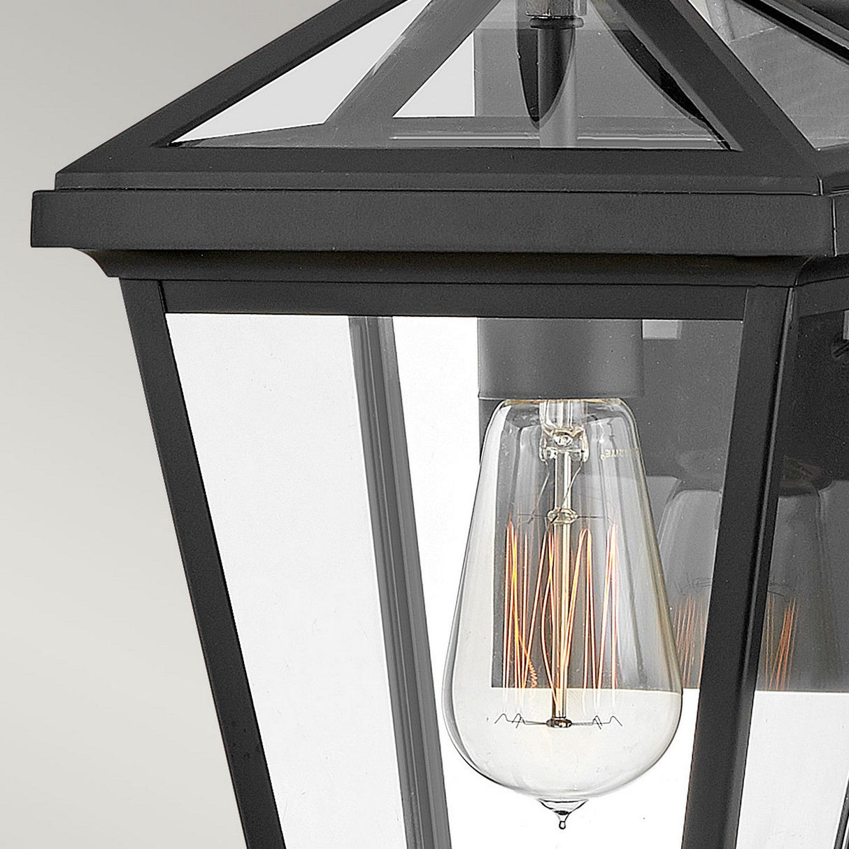 A close-up of the Palma Outdoor 1 Light Small Wall Lantern showcases its clear glass panels revealing a vintage-style Edison bulb inside. This lantern, with a sleek black finish and geometric top design, is subtly accented with hints of a pineapple motif. The unlit bulb highlights its intricate filament details.