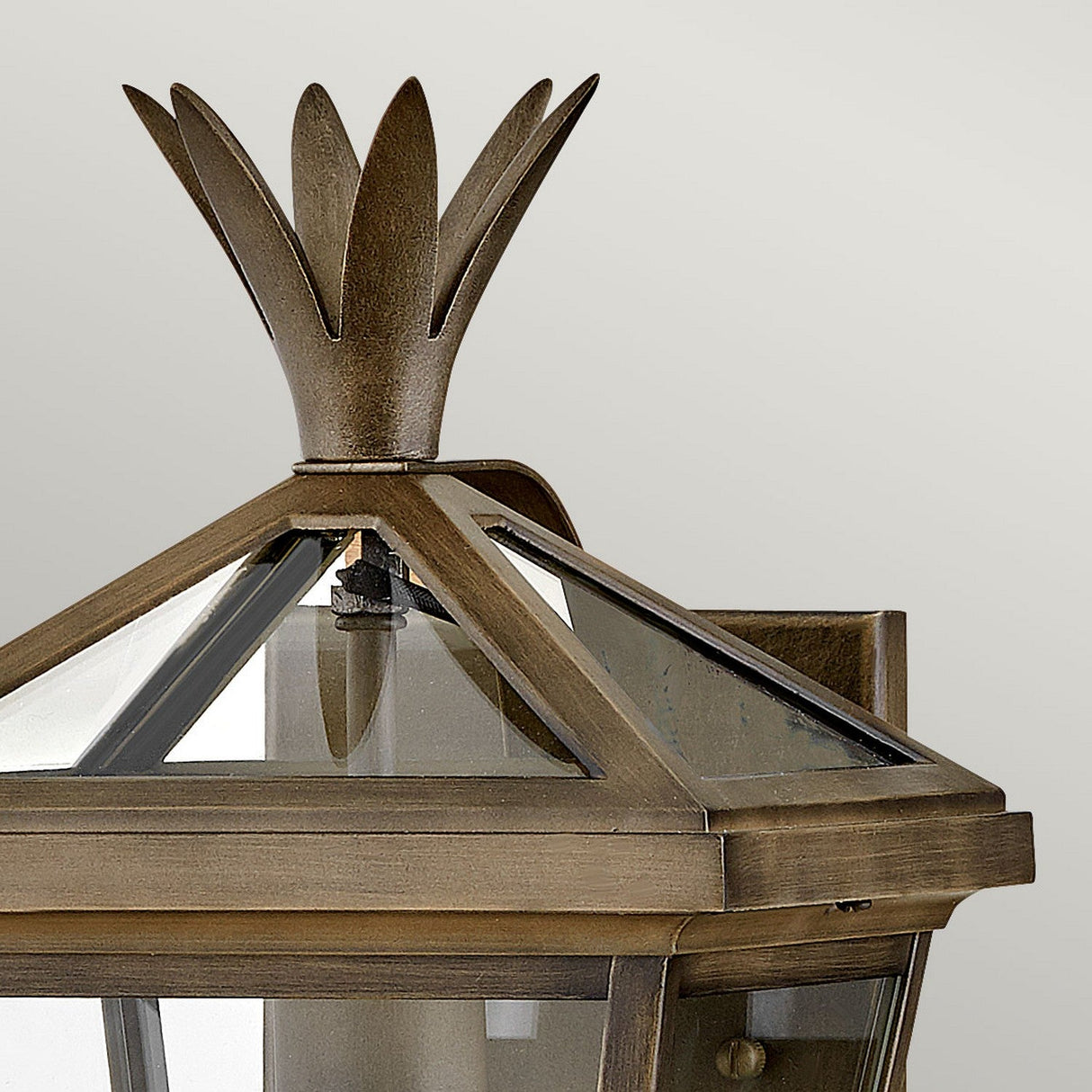 A close-up view of the Palma Outdoor 1 Light Small Wall Lantern, showcasing its triangular glass design and crown-like finial in a burnished bronze finish. The base features a subtle pineapple motif, all set against a soft, neutral background.