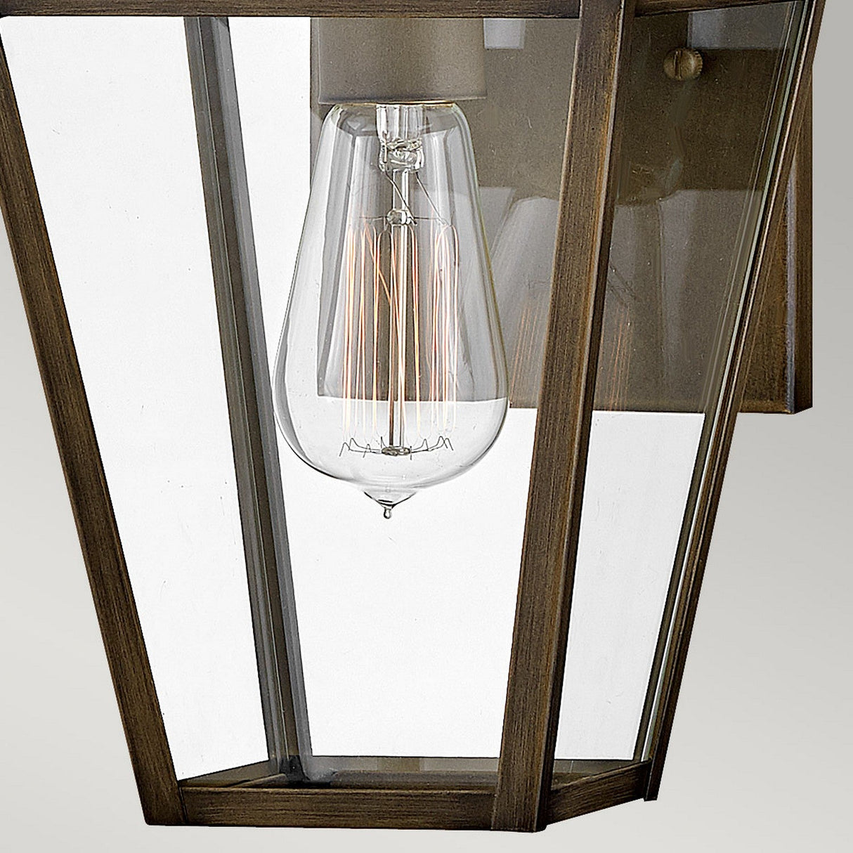 Close-up of the Palma Outdoor 1 Light Small Wall Lantern in burnished bronze, featuring a modern design with clear glass enclosure and an exposed filament light bulb encapsulated in a sleek, geometric metal frame.