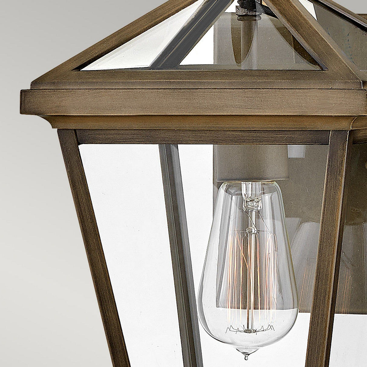 A close-up of the Palma Outdoor 1 Light Small Wall Lantern in burnished bronze showcases its vintage design with a clear glass enclosure and an exposed filament bulb. Its frame, featuring a pyramid-shaped top, radiates elegance and classic charm, while a subtle pineapple motif enhances its sophistication against the neutral background.