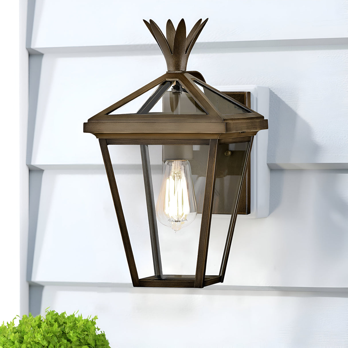 The Palma Outdoor 1 Light Small Wall Lantern in burnished bronze, featuring a geometric design with a subtle pineapple motif, adorns a light gray wall. Its glass panels showcase the visible incandescent bulb, harmonized by a small green shrub peeking in the bottom left corner.