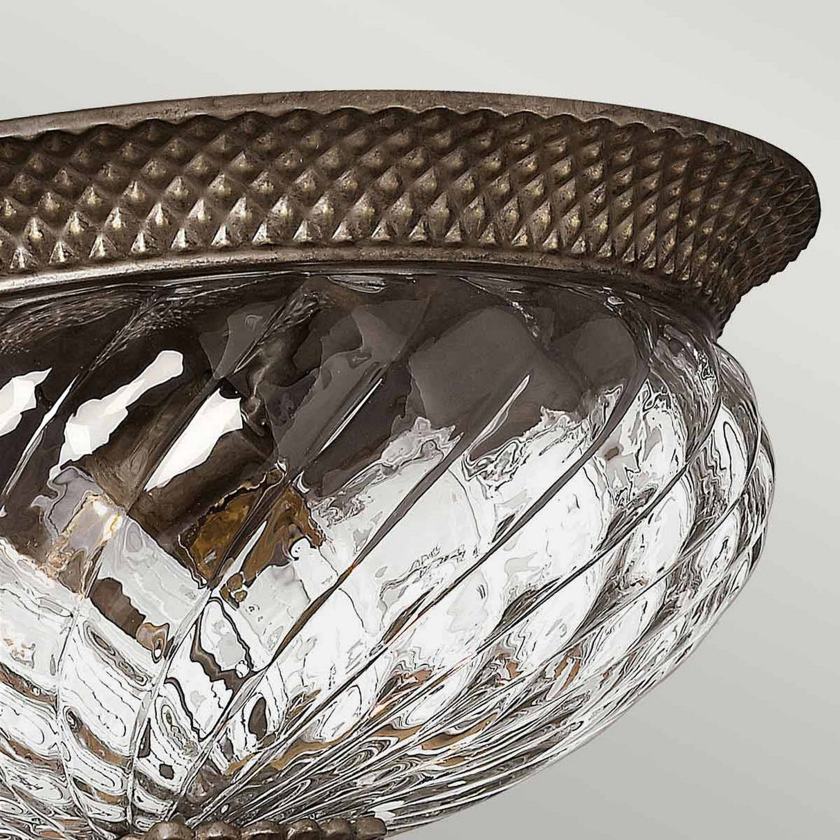 A close-up of the Plantation 3 Light Large Flush in Bronze showcases an elegant ceiling light fixture with a textured metallic rim and a clear, pineapple-shaped glass dome. The intricate geometric patterns in the design add a sophisticated touch to any decor.