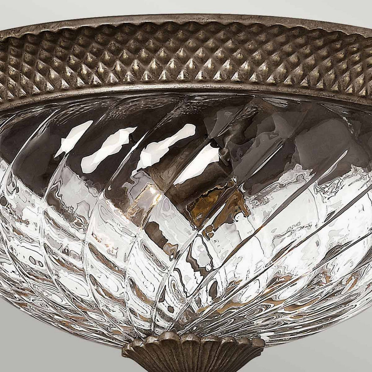 Close-up of the Plantation 3 Light Large Flush - Bronze, a decorative glass ceiling light fixture with a bronze flush mount. The swirling pineapple-shaped glass beautifully complements the metal rim's intricate diamond pattern. The soft light it casts highlights the detailed design.