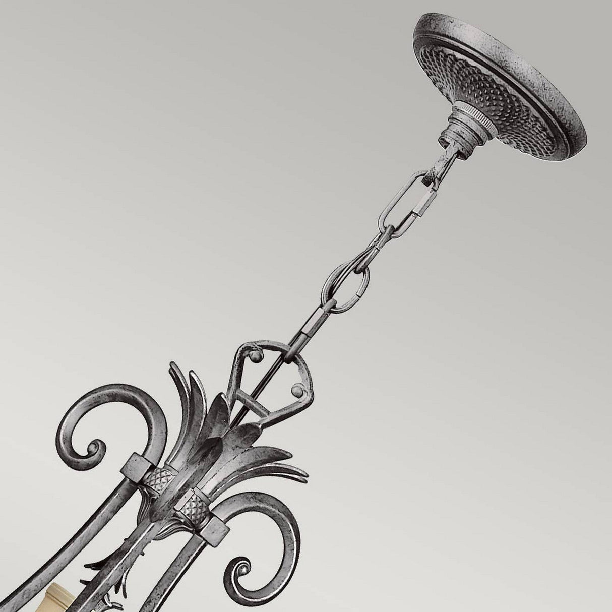 The Plantation 10 Light Chandelier showcases a close-up of its decorative metal chain and circular ceiling mount. The chain beautifully connects to a scrollwork design adorned with floral motifs. It features a dark, textured finish in Polished Antique Nickel, elegantly contrasting against a light background.