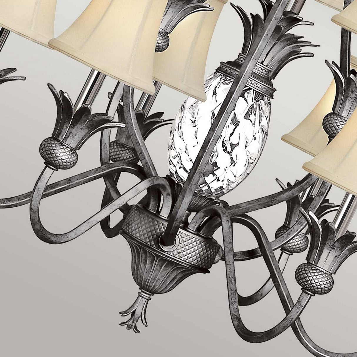 The Plantation 10 Light Chandelier in polished antique nickel is a decorative piece featuring a pineapple motif and intricate metalwork. It includes textured frosted glass shades shaped like pineapples, several gracefully curved arms with cream-colored lampshades, and adds an elegant touch to any room.