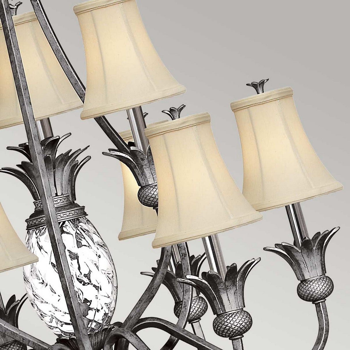 The Plantation 10 Light Chandelier in polished antique nickel features intricate metal detailing, pineapple-shaped glass shades, and cream-colored lampshades, offering an elegant blend of tropical-inspired accents against a neutral backdrop.